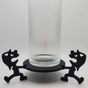 May include: A clear, ribbed glass cylinder sits on a black, circular base supported by two black, stylized figures. The figures appear to be running or dancing, adding a whimsical touch to the design. The glass is empty, showcasing its vertical ribbing.