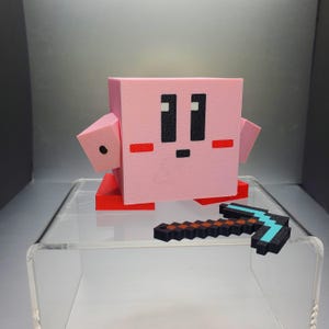May include: A pink, pixelated character figure with black eyes and a small mouth, red feet, and arms. A small, pixelated pickaxe with a black, brown, and blue design is in front of the figure. The figure is on a clear, acrylic display.