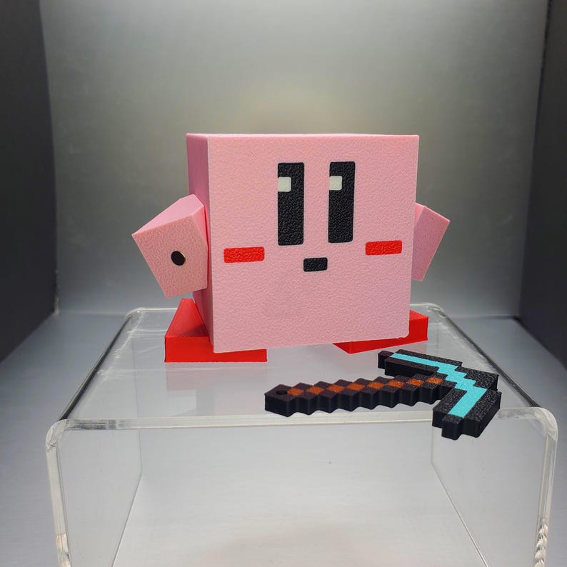 May include: A pink, pixelated character figure with black eyes and a small mouth, red feet, and arms. A small, pixelated pickaxe with a black, brown, and blue design is in front of the figure. The figure is on a clear, acrylic display.