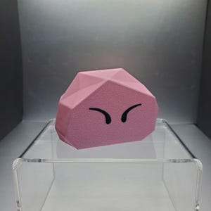 Op de afbeelding: Een roze, geometrisch gevormd decoratief object met een getextureerd oppervlak en zwarte, gebogen oogmarkeringen. Het object wordt gepresenteerd op een heldere acryl plank tegen een grijze achtergrond. Het ontwerp doet denken aan een stripfiguur.