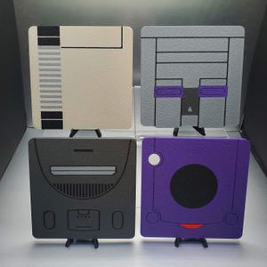 May include: Four square coasters featuring retro video game console designs. The coasters include designs inspired by the NES, Super Nintendo, Sega Saturn, and GameCube consoles. Each coaster is a different color, including beige, gray, black, and purple.