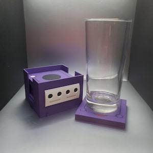 Può includere: Un set di sottobicchieri a tema GameCube viola e bianco con una scatola abbinata. Il sottobicchiere è quadrato, con un design GameCube, e un bicchiere trasparente è appoggiato sopra. La scatola è una replica in miniatura della console.