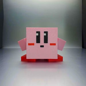 May include: A pink, cube-shaped figure with black eyes, a small black mouth, and red cheek markings. The figure has small pink arms and is mounted on a red base. The design is reminiscent of a popular video game character.