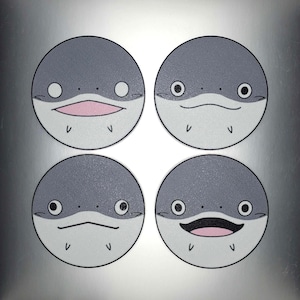 May include: Four round coasters with a cartoon fish face design. Each coaster features a gray top half and a white bottom half, with different expressions: open mouth, closed mouth, and two with a straight line mouth.
