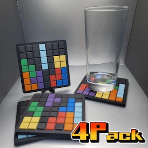 Może przedstawiać: Zestaw czterech czarnych kwadratowych podkładek z kolorowym wzorem przypominającym Tetris. Każda podkładka ma siatkę małych kwadratów w różnych kolorach, z przezroczystym szkłem na jednej. Widoczny jest tekst "4 Pack".