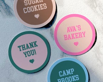 Puede incluir: Cuatro pegatinas redondas con texto y gráficos de corazón. Las pegatinas incluyen las frases "Sugar Cookies", "Ava's Bakery", "Thank You!" y "Camp Brooks". Cada pegatina tiene un esquema de color diferente, incluyendo marrón, rosa, verde y azul.