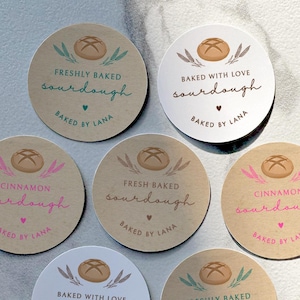 May include: A collection of round stickers with various text and designs. The stickers feature a bread illustration, the words "sourdough", "cinnamon", and "baked by Lana". Some stickers say "freshly baked" or "baked with love".