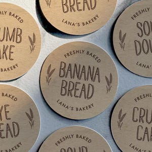 May include: Close-up of several round, brown paper labels with the text "FRESHLY BAKED" and "LANA'S BAKERY" in dark brown. The labels feature text such as "BANANA BREAD" and "SOUR DOUGH".