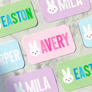 May include: A collection of pastel-colored rectangular cards with names in bold text. Each card features a cute white bunny face. The names include "Easton," "Avery," and "Mila." The cards are on a light gray surface.