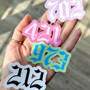 May include: A hand holding several die-cut stickers with area code numbers. The stickers are in various colors, including pink, green, and purple, with white outlines. The numbers are in a stylized font. The text "littlemagicprints" is at the bottom.