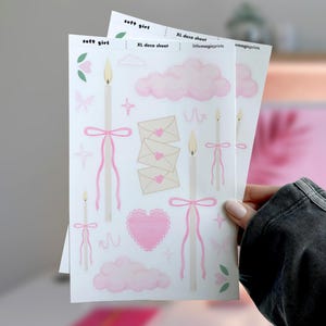 Puede incluir: Dos hojas XL decorativas con un tema de chica suave. Las hojas presentan ilustraciones de velas con lazos rosas, nubes rosas, corazones, sobres y el texto "soft girl". Las hojas se sostienen en la mano de una persona.