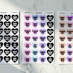 May include: Three sheets of heart-shaped stickers with days of the week written on them. The first sheet has black hearts with white text. The second sheet has pastel pink, purple, and blue hearts with black text. The third sheet has peach and orange hearts with black text.