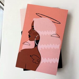May include: A pink and peach notebook with a black and white illustration of a woman with pink hair and a halo. The woman has brown skin and is wearing a hoop earring. The background is a peach color with white clouds.