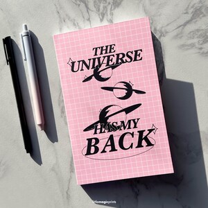 May include: Pink notebook with a grid pattern and black text that reads "The Universe Has My Back". The text is surrounded by black stylised planets.