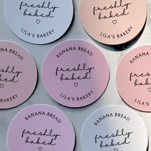 May include: Six round stickers with a pink and white gradient. The stickers say "Banana Bread freshly baked" with a heart symbol and "Lila's Bakery" in black text.