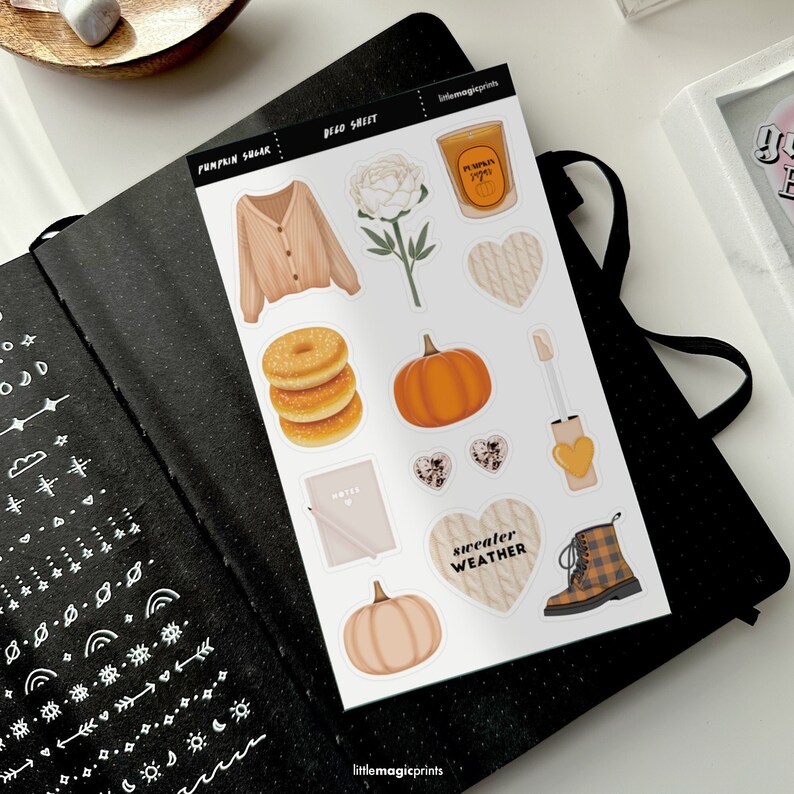 May include: A sticker sheet with fall-themed illustrations, including a pumpkin, donuts, a sweater, and a candle. The sheet is labeled "PUMPKIN SHEAR" and "DECO SHEET" with the text "sweater weather". The stickers are on a black notebook.