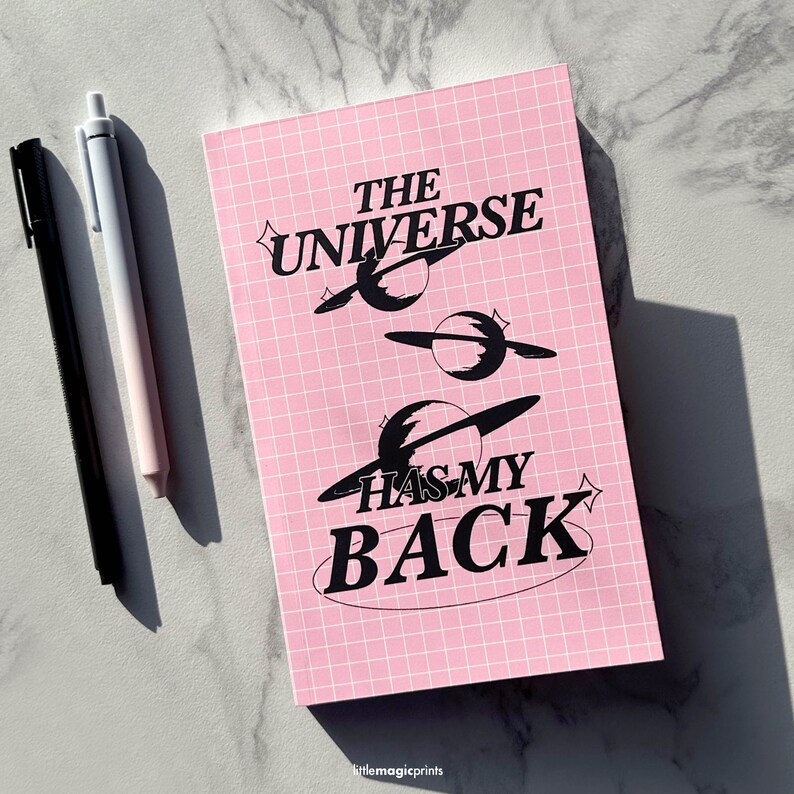 May include: Pink notebook with a grid pattern and black text that reads "The Universe Has My Back". The text is surrounded by black stylised planets.