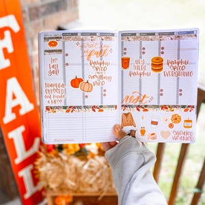 May include: Open planner with fall-themed stickers and handwritten text. The planner includes phrases like "It's Fall Y'all!" and "Pumpkin Spice Everything." Decorative elements include pumpkins, leaves, and coffee cups. The planner is open to the week of September 21-27.