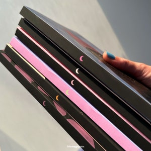 May include: A stack of five black notebooks with pink and purple accents. Each notebook has a crescent moon design on the spine.