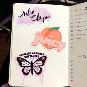 May include: A planner page with three stickers. The first sticker is a pink cloud with the text "relax and let go" in black. The second sticker is a peach with a heart on it. The third sticker is a pink butterfly with black wings and the text "socially awkward butterfly" in black.