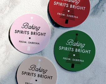 May include: Four round stickers with a Christmas tree design. Each sticker has the text "Baking Spirits Bright" and "From: Sabrina" in black text on a colored background. The stickers are pink, red, green, and beige.