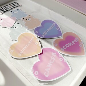 May include: A set of 5 heart-shaped stickers with zodiac signs. The stickers are in shades of pink, purple, blue, and orange. The zodiac signs are Sagittarius, Aquarius, Cancer, Scorpio.
