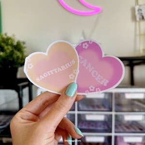 May include: Two heart-shaped stickers with a gradient design. The sticker on the left is peach and pink with the text "SAGITTARIUS" in white. The sticker on the right is pink with the text "CANCER" in white.