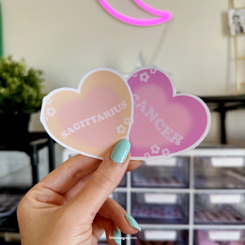 May include: Two heart-shaped stickers with a gradient design. The sticker on the left is peach and pink with the text "SAGITTARIUS" in white. The sticker on the right is pink with the text "CANCER" in white.