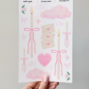 Puede incluir: Una hoja de pegatinas con tem&aacute;tica de chica suave. Las pegatinas incluyen velas con lazos rosas, nubes rosas, sobres con corazones, mariposas y flores. El texto "soft girl" y "XL deco sheet" son visibles.