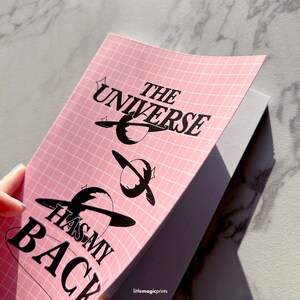 May include: A pink notebook with a grid pattern and the text "The Universe Has My Back" in black. The notebook is open to reveal a blank page with a dotted pattern.