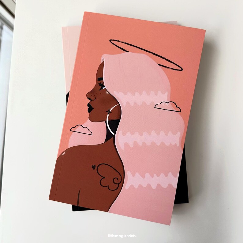 May include: A pink and peach notebook with a black and white illustration of a woman with pink hair and a halo. The woman has brown skin and is wearing a hoop earring. The background is a peach color with white clouds.