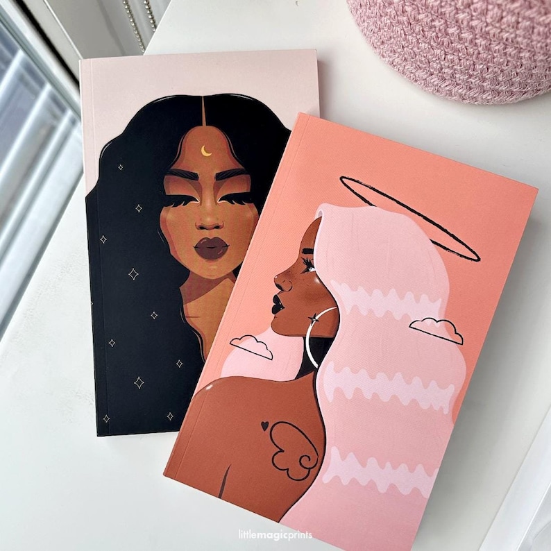 May include: Two illustrated notebooks with black and pink covers. The notebook on the left features a black woman with a crescent moon above her head. The notebook on the right features a pink-haired woman with a halo above her head. Both notebooks have the text "littlemagicprints" at the bottom.