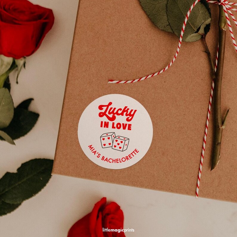 May include: A round white sticker with red text that reads "Lucky in Love" and "Mia's Bachelorette". The sticker features dice illustrations. A red rose and a brown box are also visible.