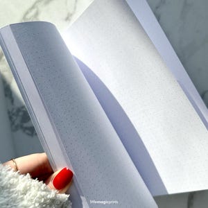 May include: An open notebook with white pages and a dotted grid pattern. The notebook is lying flat on a white surface.