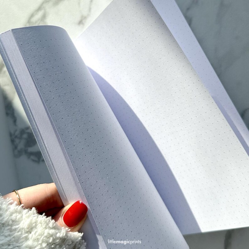 May include: An open notebook with white pages and a dotted grid pattern. The notebook is lying flat on a white surface.