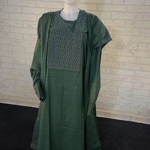 Pode incluir: Um traje tradicional masculino verde com um padr&atilde;o geom&eacute;trico no peito. O traje inclui uma t&uacute;nica longa, cal&ccedil;as e um chap&eacute;u.