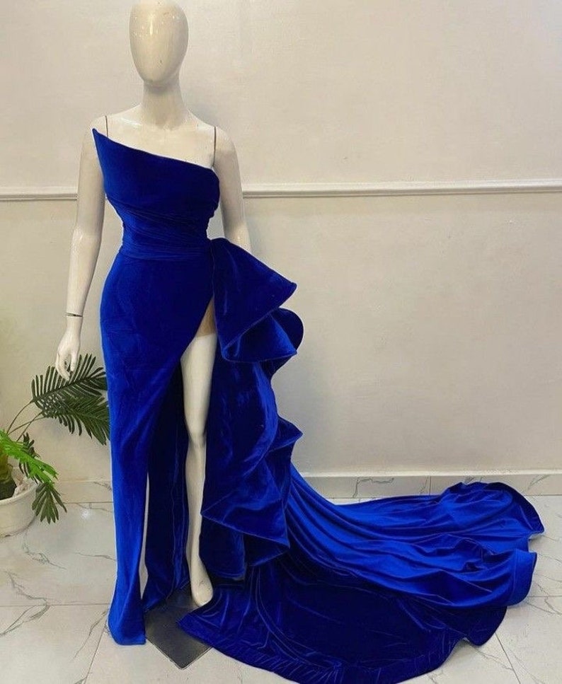 Puede incluir: Un vestido azul real de un solo hombro con una abertura alta y una cola dram&aacute;tica y fluida. El vestido presenta un gran acento con volantes en la cintura y se exhibe en un maniqu&iacute;. La tela parece ser terciopelo suave.
