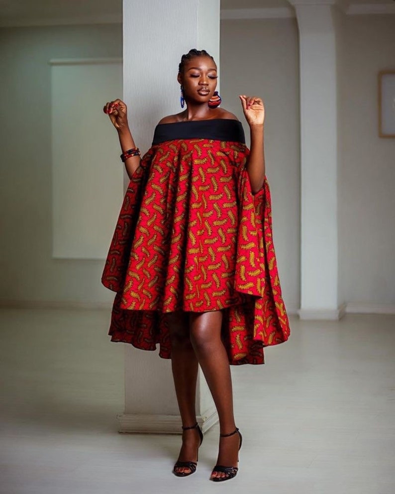 Pode incluir: Um vestido vermelho e dourado com decote ombro a ombro e uma faixa preta na cintura. O vestido tem uma bainha alta-baixa e &eacute; usado por uma pessoa de pele escura.