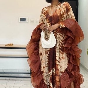 May include: A woman wearing a brown and white tie-dye maxi dress with a brown ruffled overlay. She is carrying a white beaded bag and wearing sunglasses.
