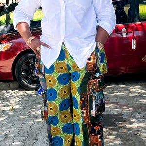 May include: A person wearing a white button-down shirt and patterned pants with blue, yellow, and green circles and geometric shapes. The pants have a wide leg and a unique design.