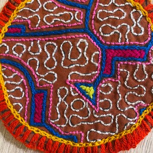 Op de afbeelding: Een ronde stoffen onderzetter met een bruine basis en gedetailleerd handborduurwerk in wit, blauw, roze en magenta garen. Het ontwerp bestaat uit abstracte, kronkelende lijnen en geometrische vormen, afgewerkt met een dikke oranje en gele geborduurde rand.
