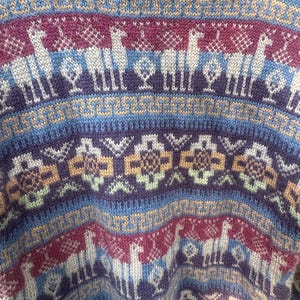 May include: A knit sweater featuring a repeating pattern of llamas, geometric shapes, and stylized trees. The color palette includes shades of deep purple, light blue, cream, and muted yellow, arranged in horizontal bands across the fabric surface.