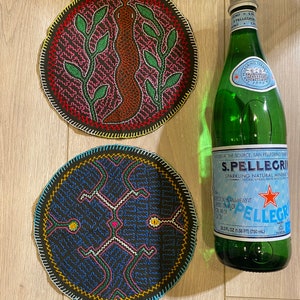 Puede incluir: Dos posavasos circulares de tela con bordados intrincados. El posavasos superior muestra una serpiente marrón flanqueada por hojas verdes sobre fondo negro. El inferior presenta un patrón geométrico azul y rosa. Se muestra una botella de 750 ml.