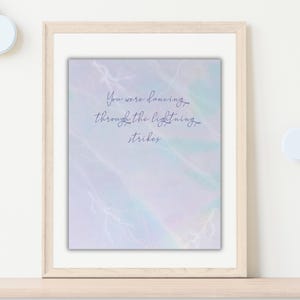 K&ouml;nnte beinhalten: Gerahmter Druck mit dem Schriftzug "You were dancing through the lightning strikes" in zarter Schrift. Der Hintergrund hat einen weichen Aquarell-Effekt mit Pastellt&ouml;nen von Blau, Lila und Gr&uuml;n, mit wei&szlig;en Blitzillustrationen.