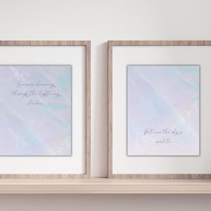 Könnte beinhalten: Zwei gerahmte Kunstdrucke mit inspirierenden Zitaten. Die Drucke zeigen einen hellblauen und lila Aquarellhintergrund mit handgeschriebenem Text. Die Rahmen sind hellbraun und stehen auf einem Holzregal. Ein Druck lautet: "You were dancing through the lightning strikes."