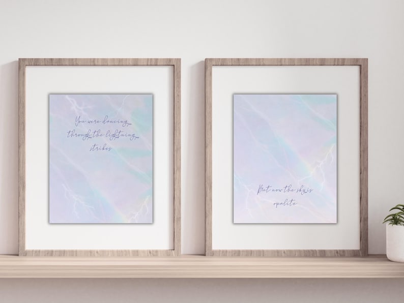 K&ouml;nnte beinhalten: Zwei gerahmte Kunstdrucke mit inspirierenden Zitaten. Die Drucke zeigen einen hellblauen und lila Aquarellhintergrund mit handgeschriebenem Text. Die Rahmen sind hellbraun und stehen auf einem Holzregal. Ein Druck lautet: "You were dancing through the lightning strikes."