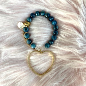 May include: A blue gemstone bracelet with a gold heart charm. The bracelet is made of round beads and has a gold clasp. The heart charm is open and has a delicate design.