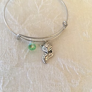 May include: Silver-tone adjustable bangle bracelet with a fox charm and a green crystal bead. The fox charm has detailed features. The bracelet is displayed on a white, textured surface.