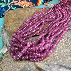May include: A strand of pink gemstone beads, with a glossy finish, arranged in a pile on a textured grey surface.