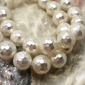 Könnte beinhalten: Eine Strähne weißer, facettierter, runder Perlen mit irisierendem Schimmer. Die Perlen sind kreisförmig angeordnet und liegen auf einer Muschel.